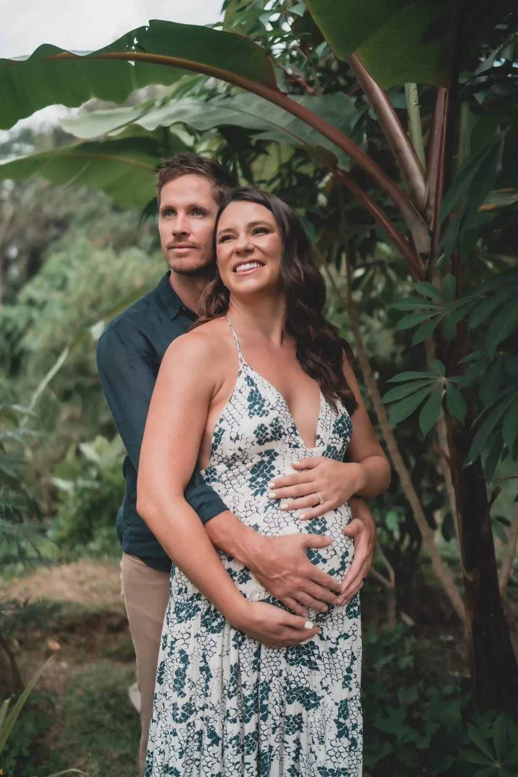 An expectant couple standing together under banana palms, her in a floral dress, his hand on her belly