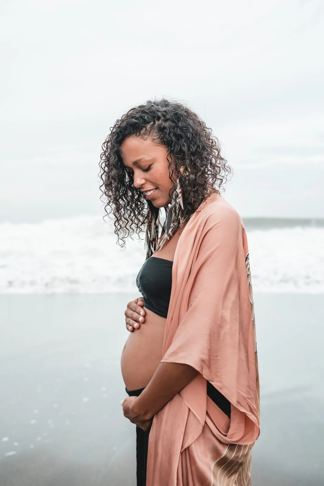A pregnant woman in a peach kimono on the beach, smiling down at her belly, the sea behind her