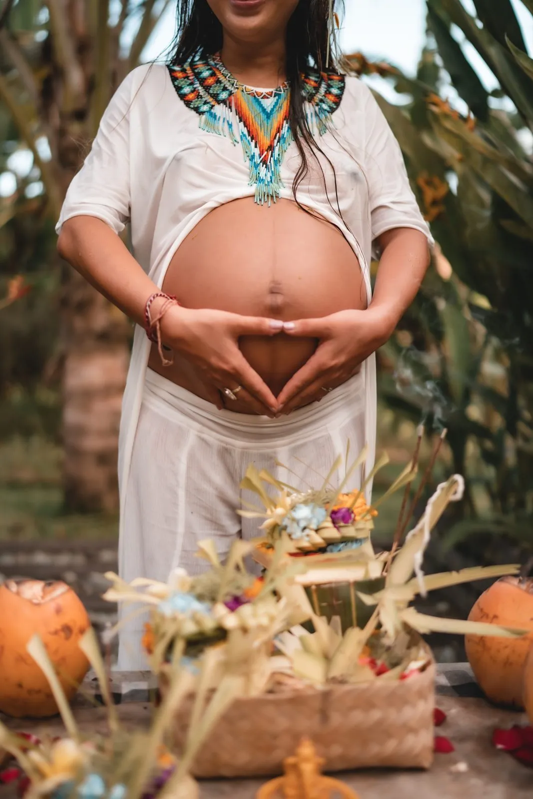 Hands cradling a bare pregnant belly at a blessingway in Bali, offerings and marigolds on the mat below