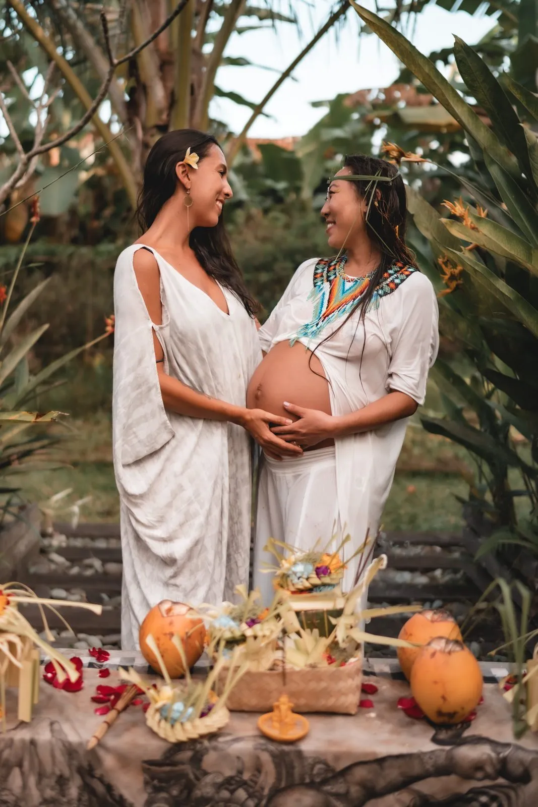 Two women in white at a blessingway ceremony among palms, one pregnant with a hand on her belly