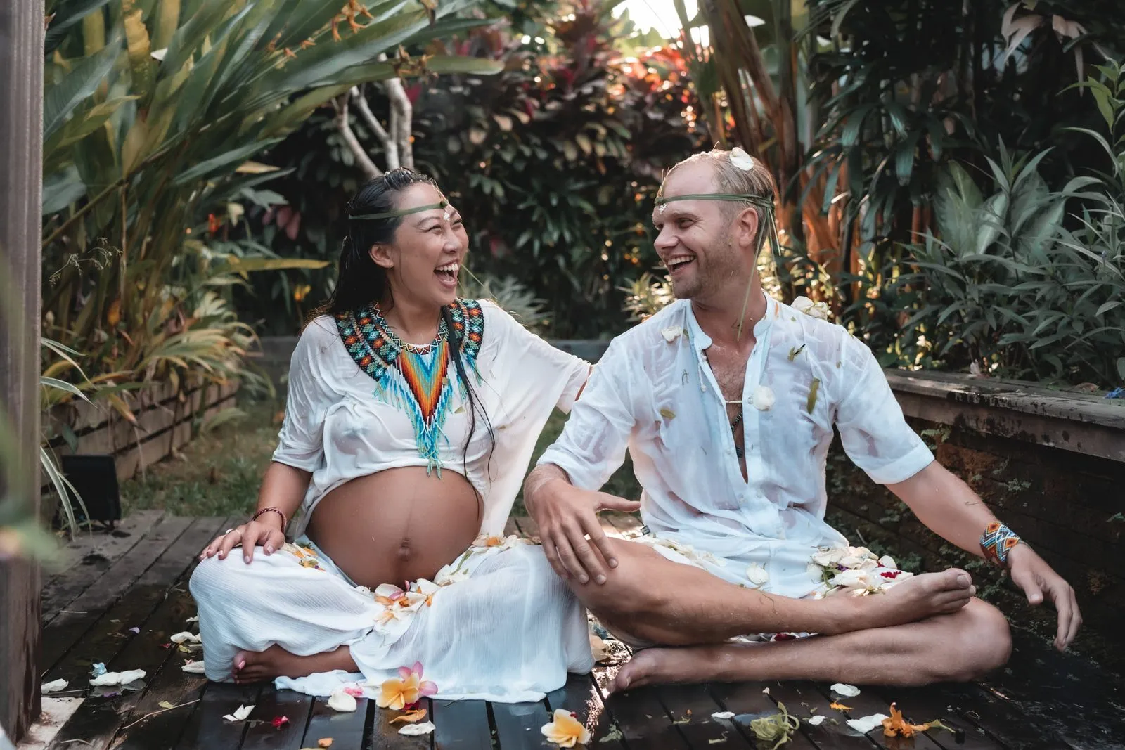 An expectant couple in white sitting cross-legged among petals at their blessingway, laughing