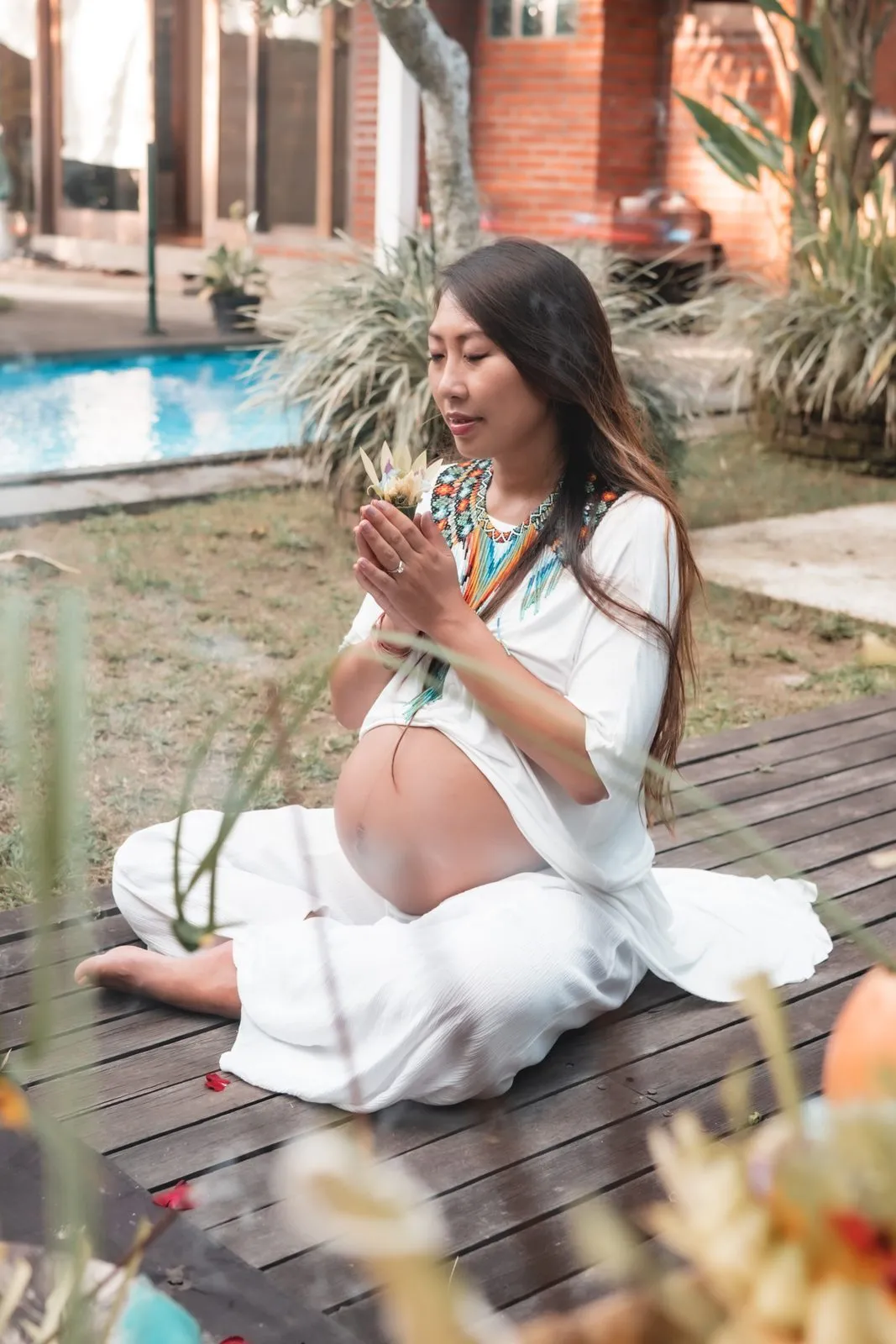 A pregnant woman in white sitting on a wooden deck by a pool, hands in prayer, eyes closed