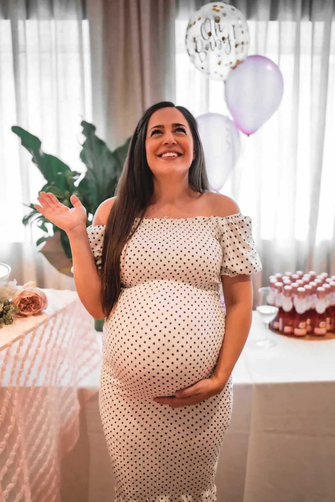 A pregnant woman in a polka dot dress laughing at her baby shower, a dessert table behind her