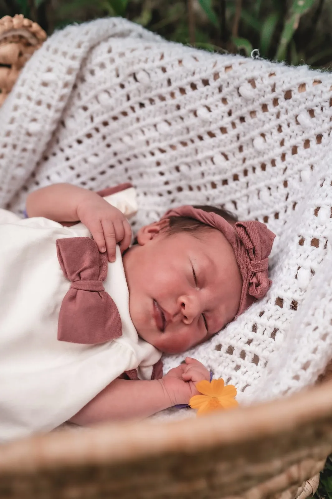 A newborn asleep in a basket under a crochet blanket, a pink bow in her hair