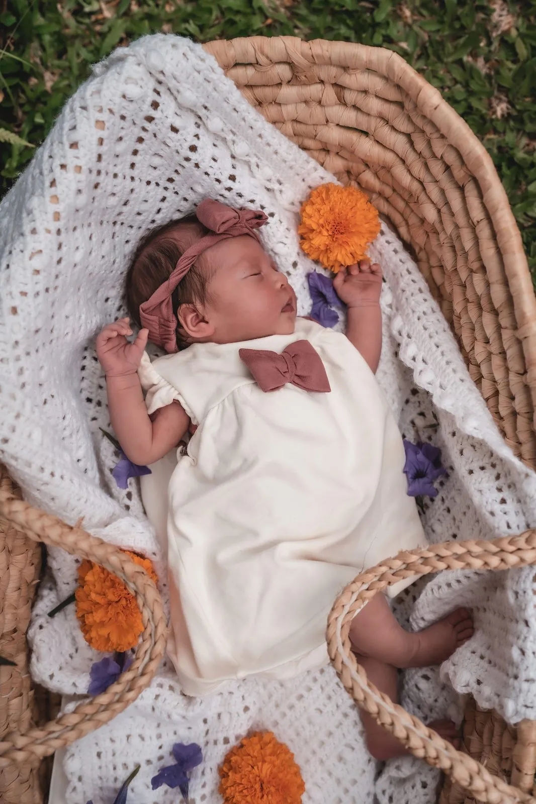 A newborn in white asleep in a woven basket with marigolds tucked around her