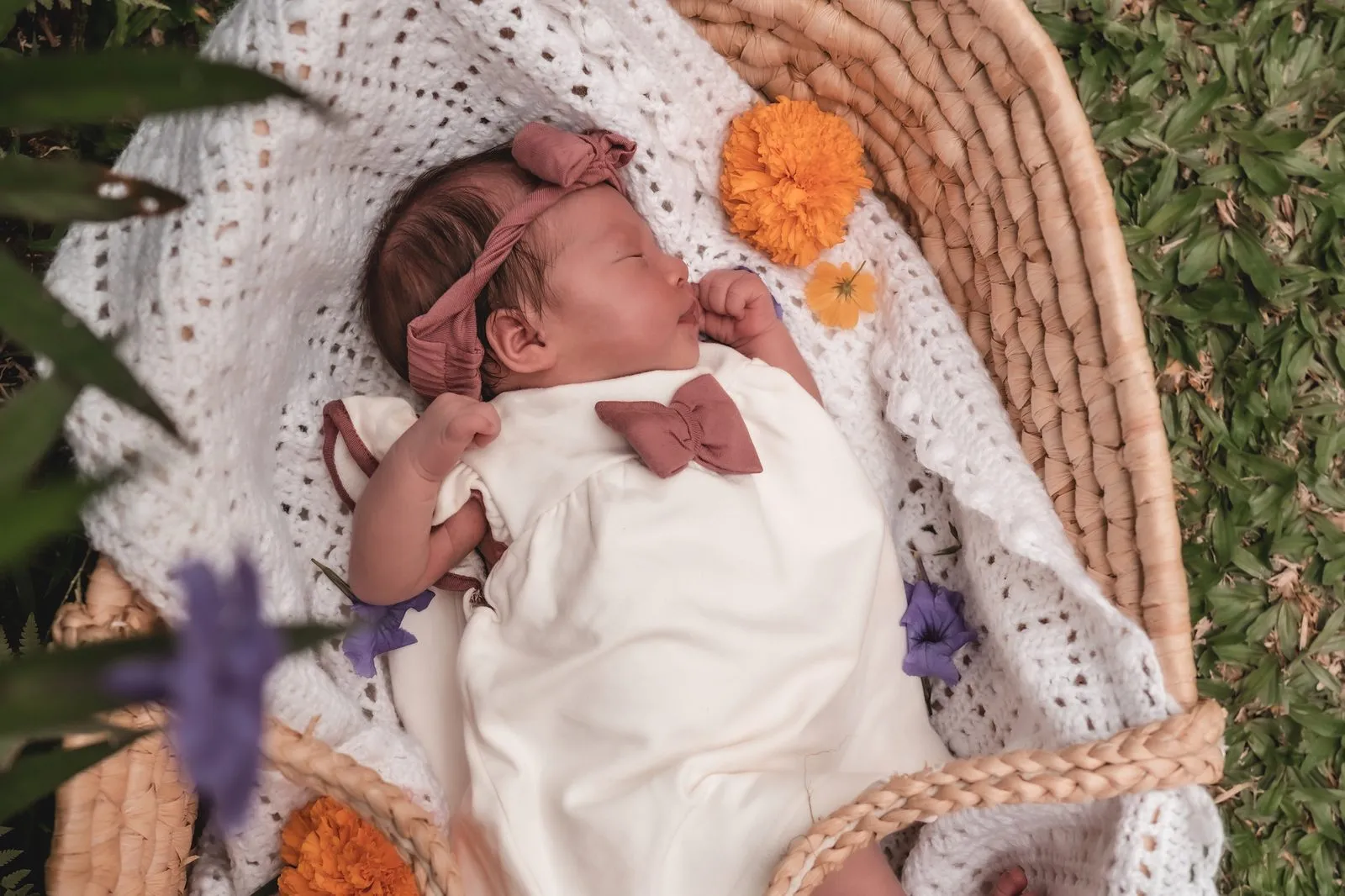 A newborn with a pink bow asleep in a basket on the grass, marigolds and lavender beside her