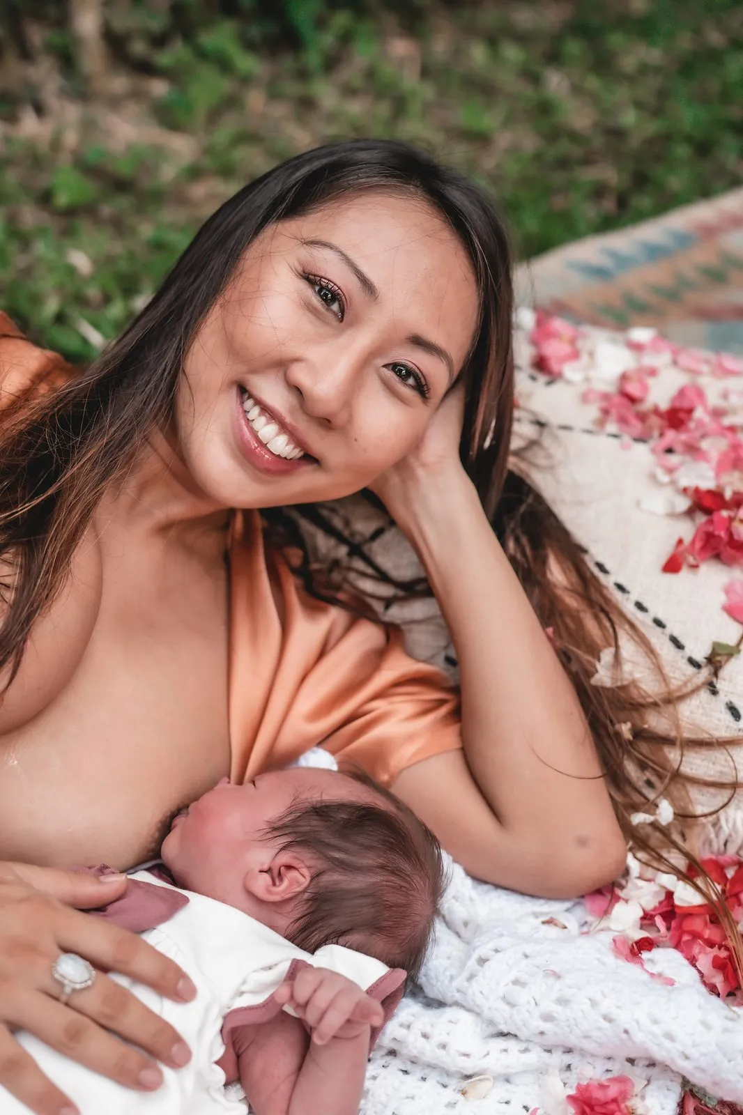 A mother in a peach robe smiling to camera as she breastfeeds her newborn