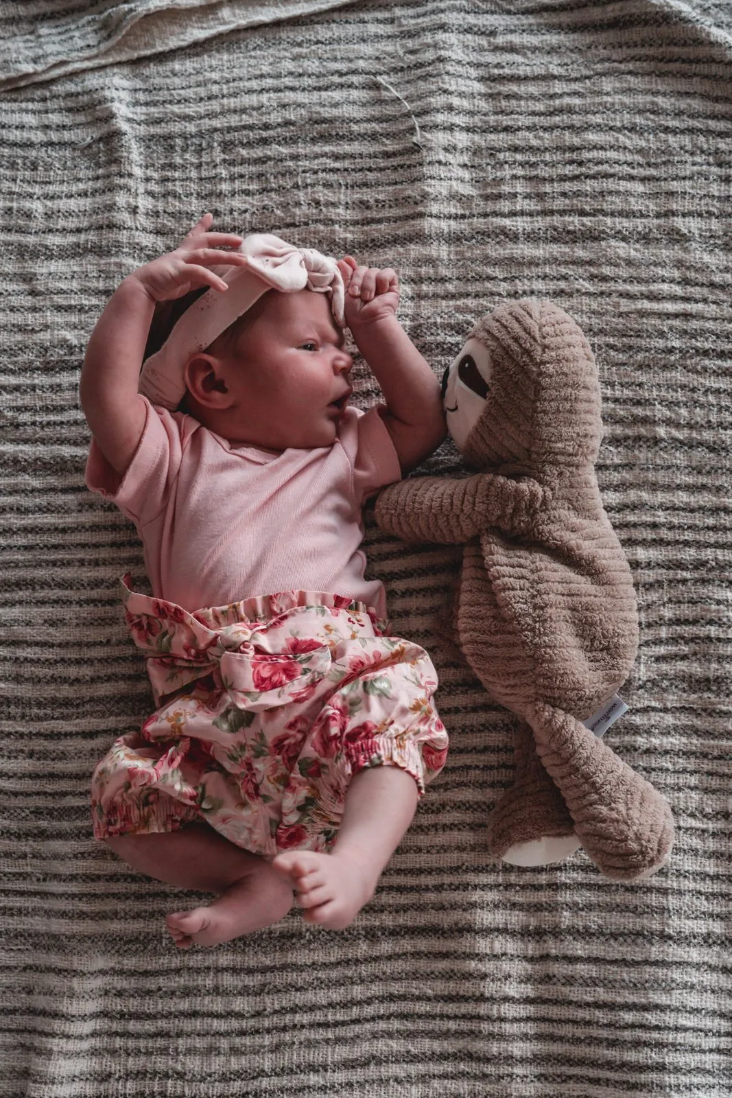 A newborn in a pink bow and floral shorts lying on a striped rug beside a knitted bear
