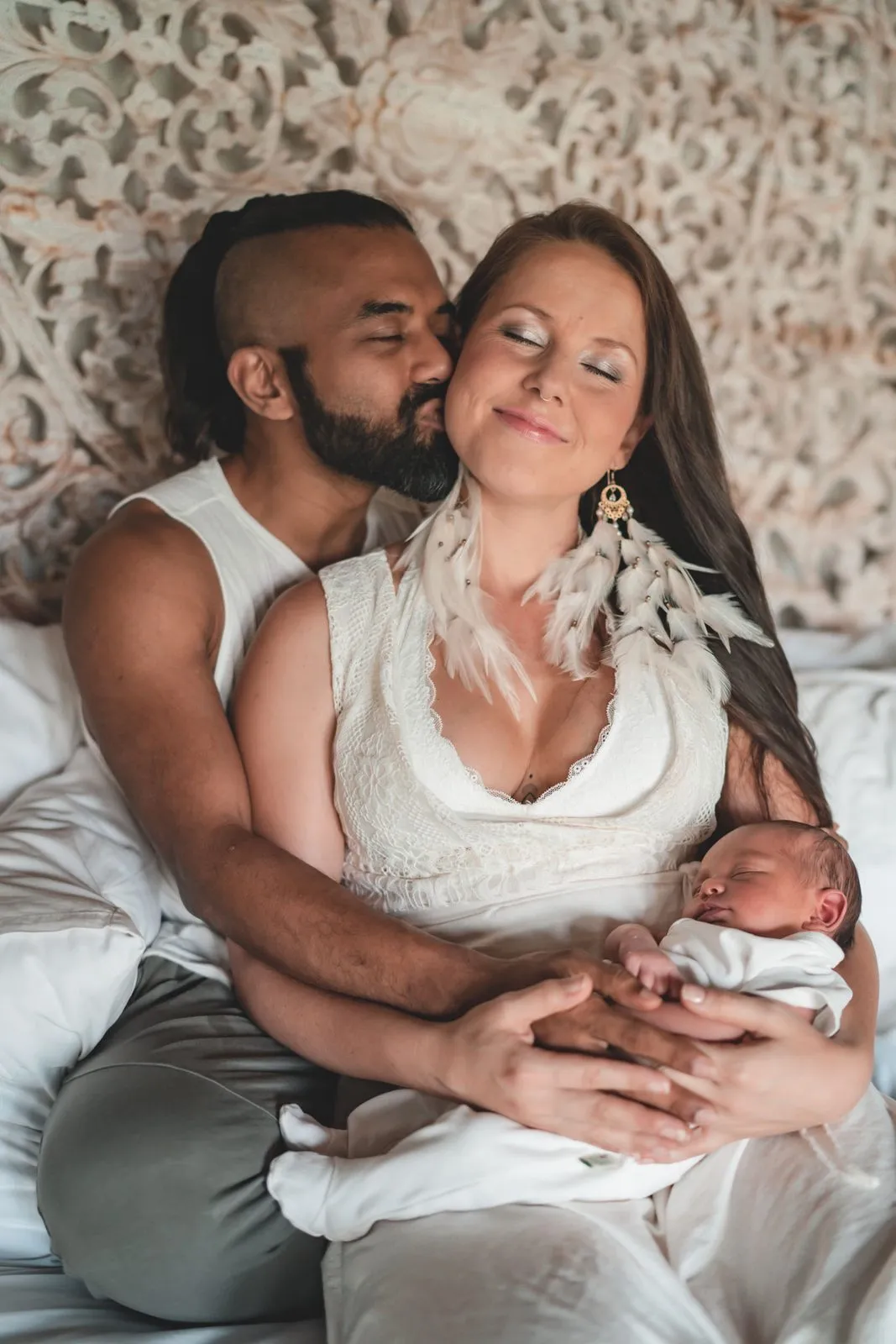 A couple on a bed holding their newborn between them, his lips to her temple