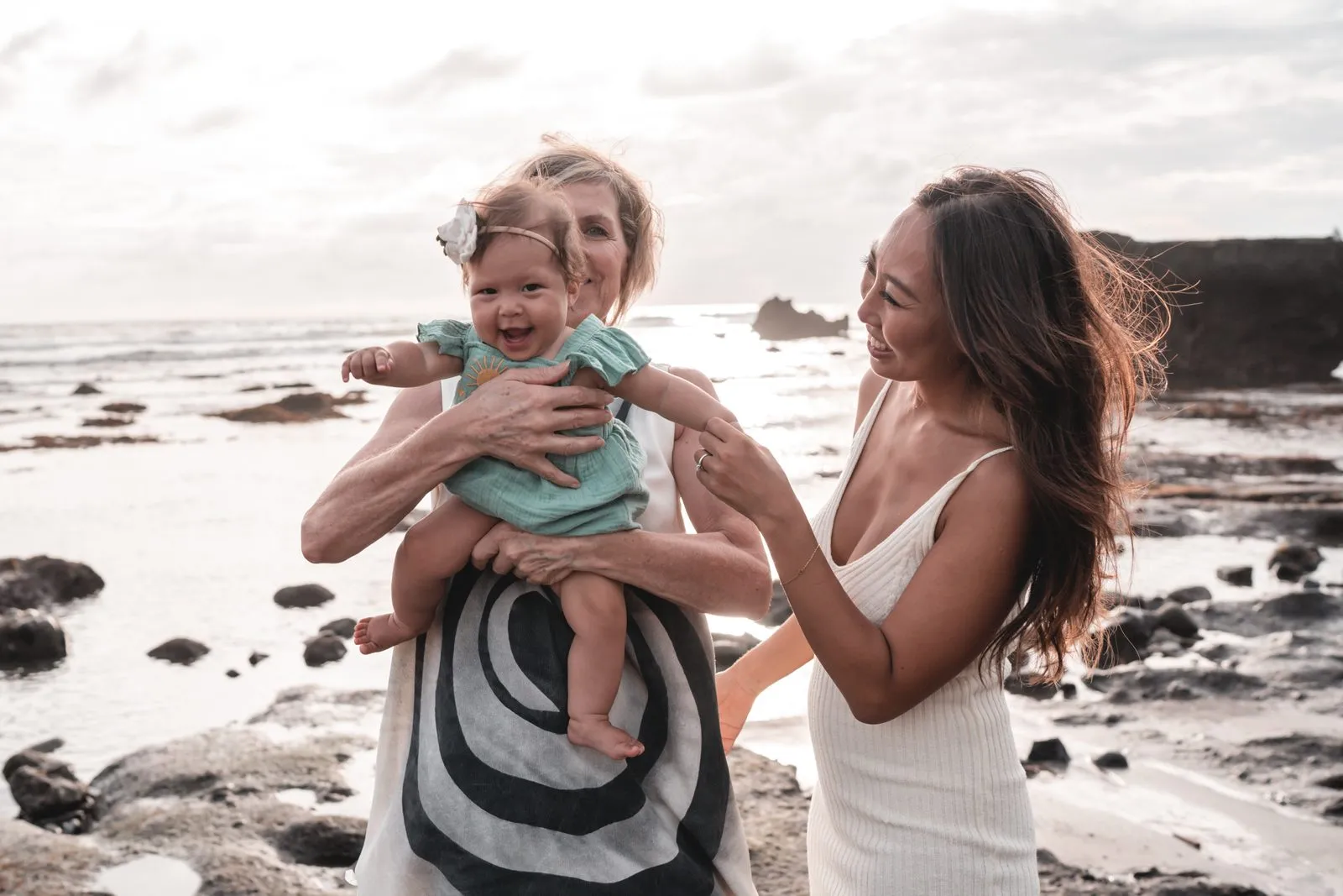 A mother and grandmother playing with a laughing baby on the rocks by the sea