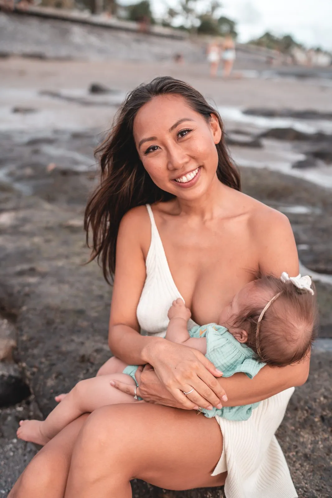 A mother in white sitting on the rocks at the beach breastfeeding her baby, smiling to camera