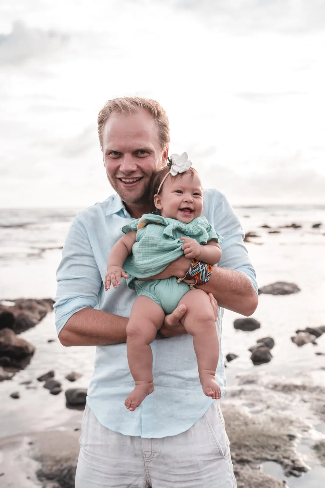 A father in a pale shirt holding his baby on the rocks at the edge of the sea