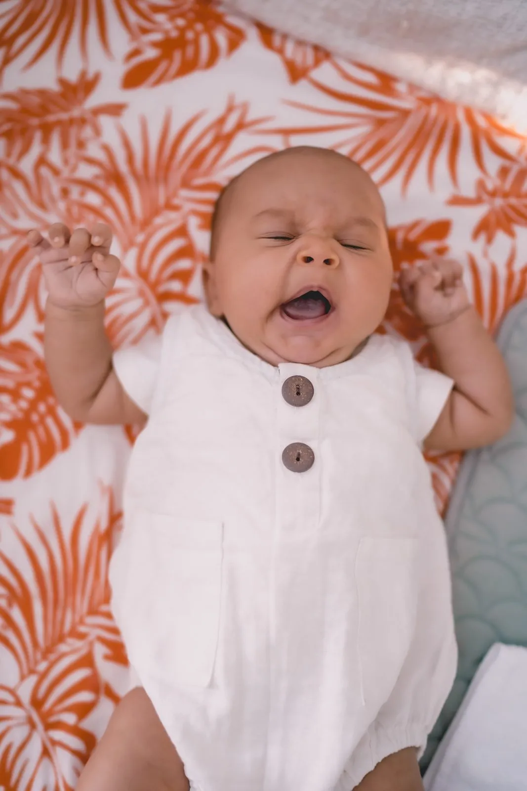 A baby in a white romper yawning on a palm-print blanket