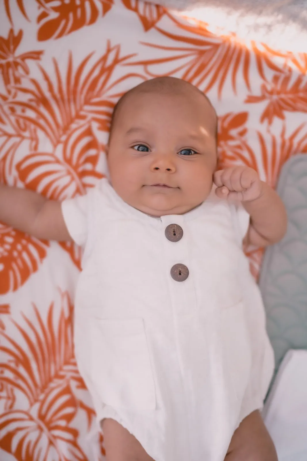 A baby in a white romper lying on an orange palm-print blanket, looking up