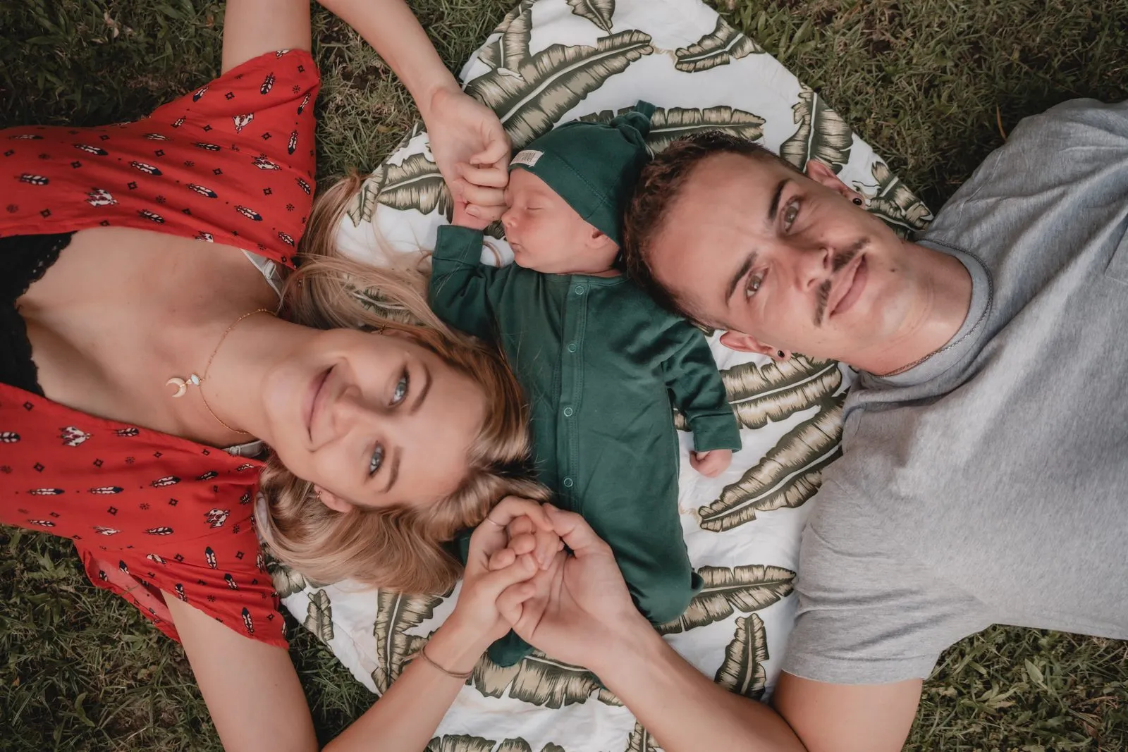 A family of three lying on the grass with their heads together, the baby held up between the parents