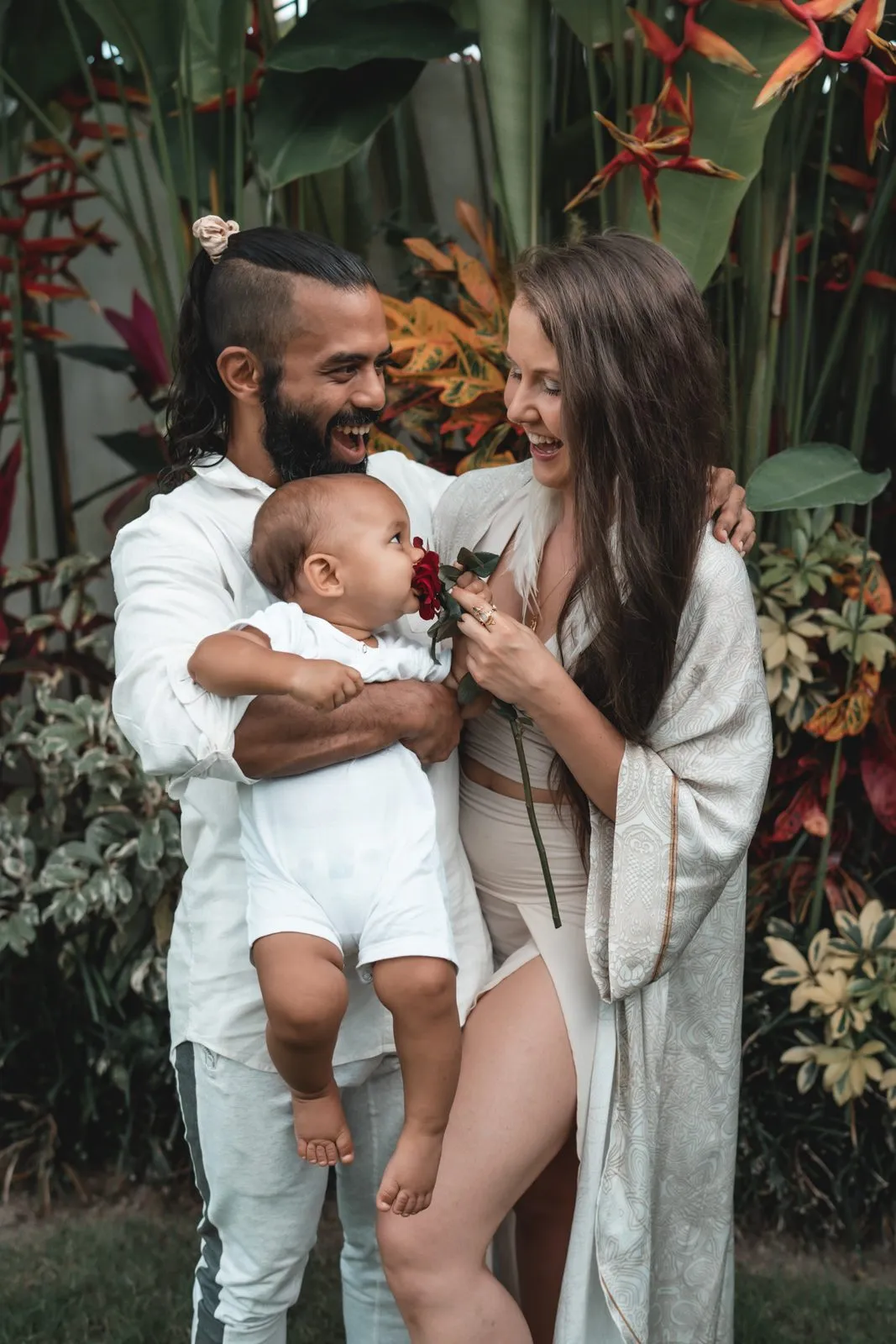 A father holding a baby while the mother leans in laughing, in a garden of red heliconia
