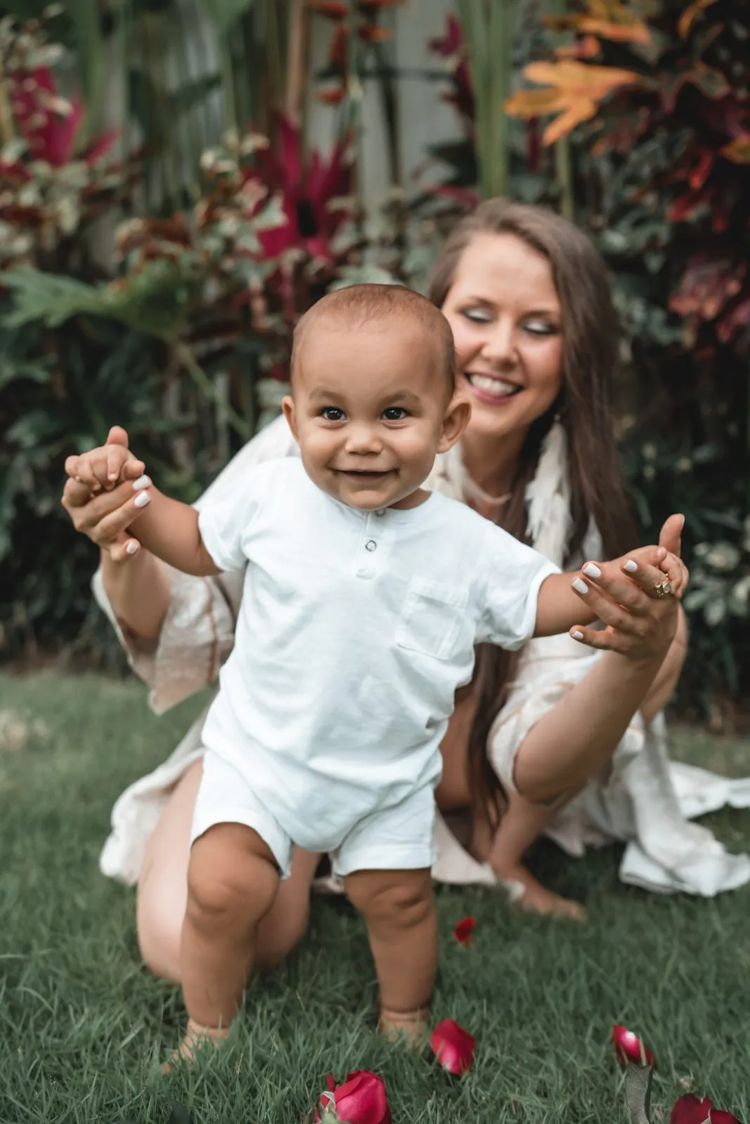 A laughing baby in white held standing on the grass by his mother, tropical leaves behind them