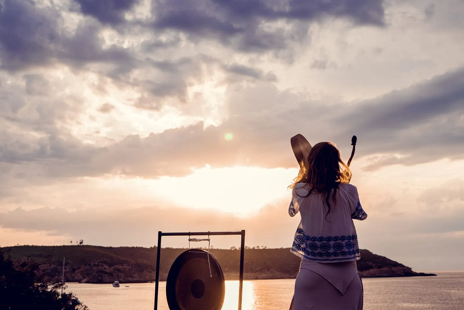 A sound healer playing a gong on the shore at sunset, the sun breaking through cloud over the sea