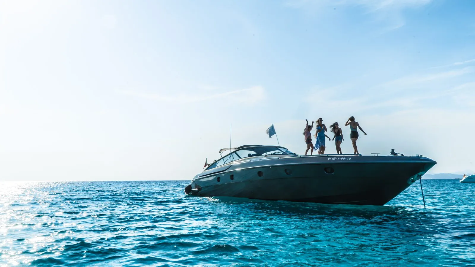 Four people dancing on the bow of a motor yacht anchored in turquoise water off Ibiza, for a boat charter brand film