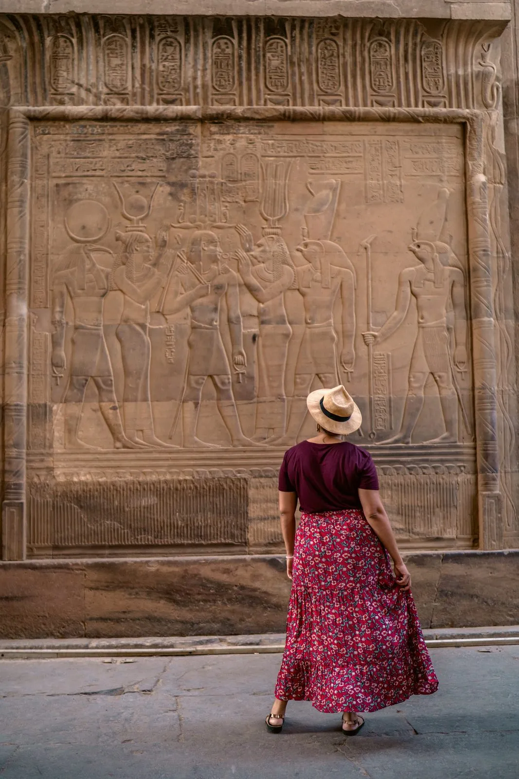 A woman in a hat and a floral skirt looking up at a wall of carved reliefs at a temple in Egypt