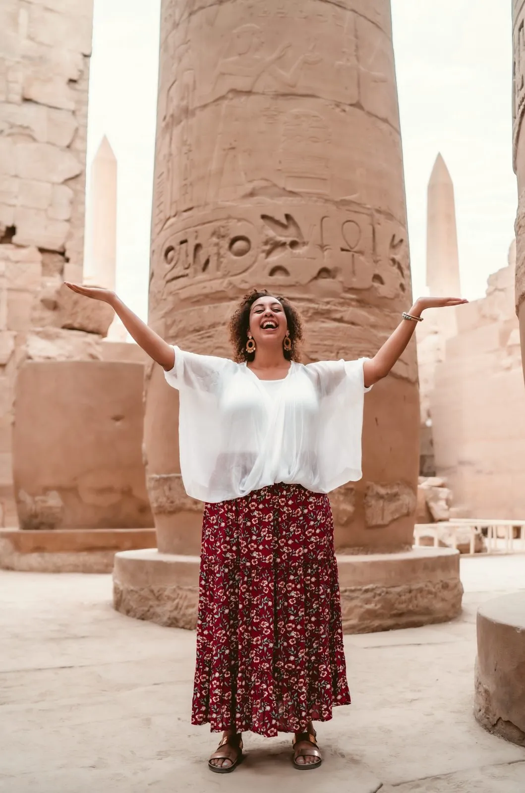 A woman in a white blouse and floral skirt with her arms open beneath the great columns of Karnak, Luxor