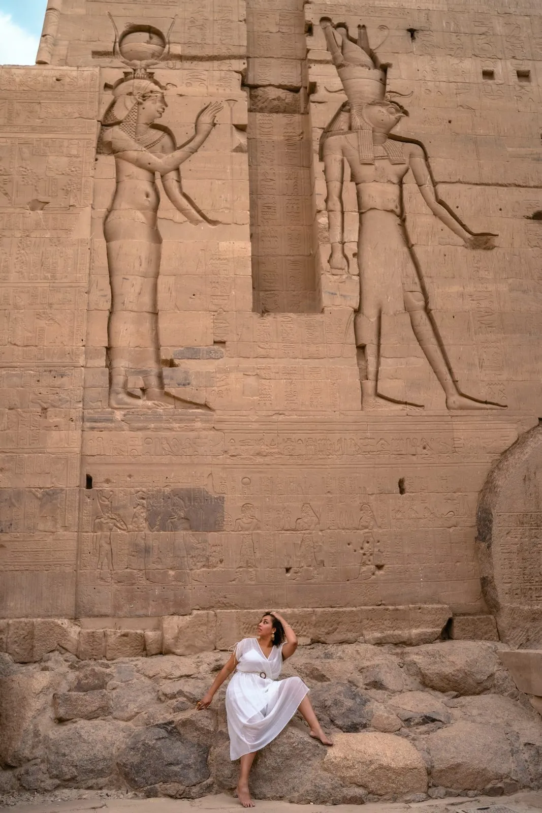 A woman in white sitting at the foot of a temple wall carved with gods, Egypt