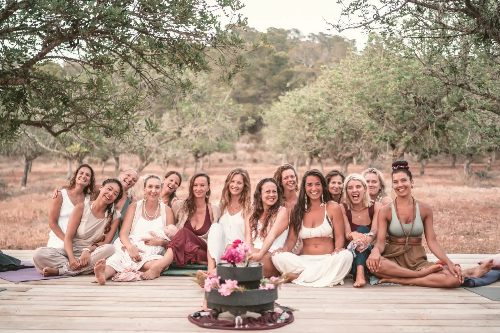 A retreat group of women sitting together on a wooden deck under the trees, flowers in the centre