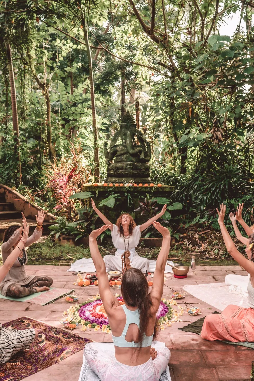 A circle of women with arms raised around a mandala of flowers under the jungle canopy, a stone Buddha above them, Bali
