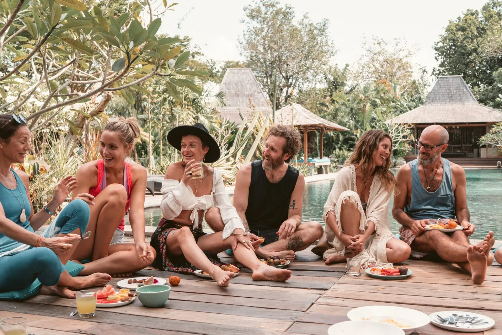 A retreat group laughing together over a shared meal on a deck by a villa pool, Bali