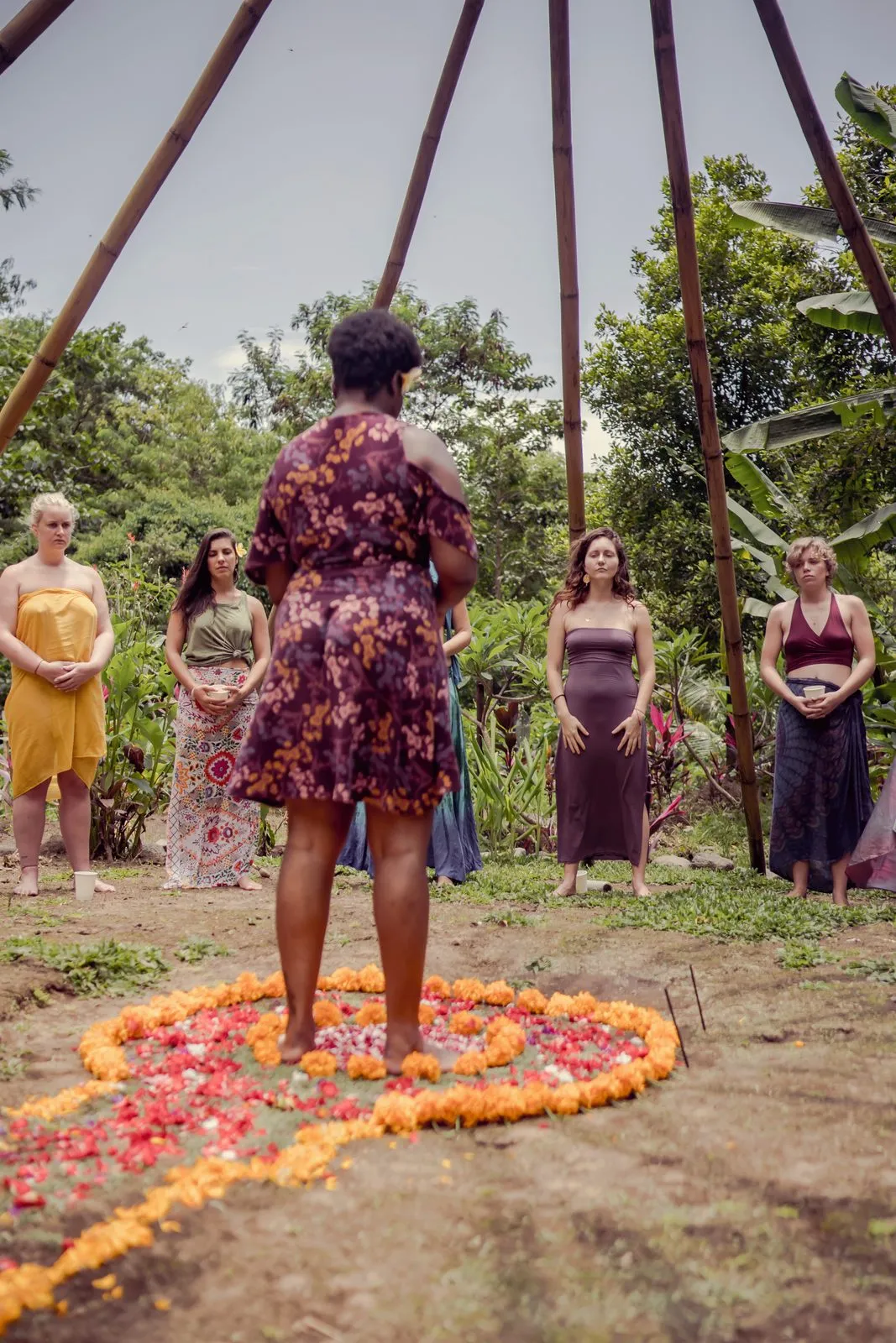 A woman in a floral dress leading a circle of women around a mandala of marigold petals under a bamboo structure