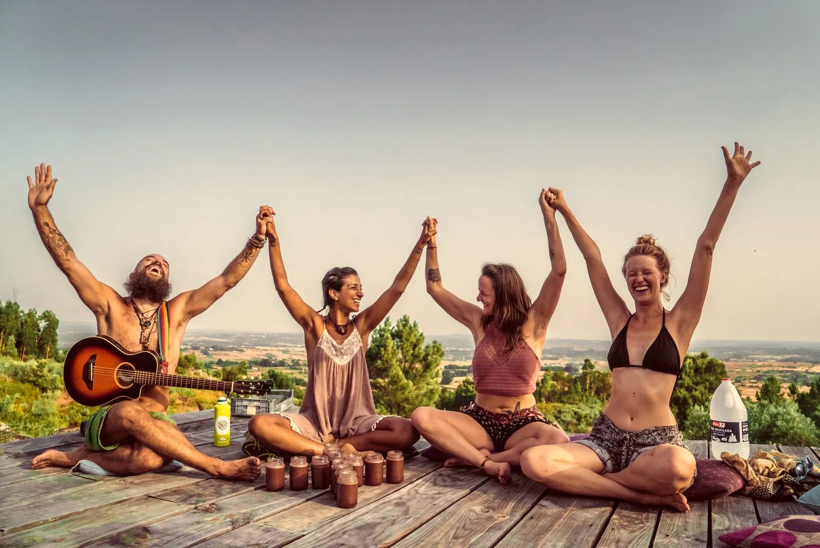 A group on a rooftop deck at sunset with their hands joined and raised, a guitar beside them