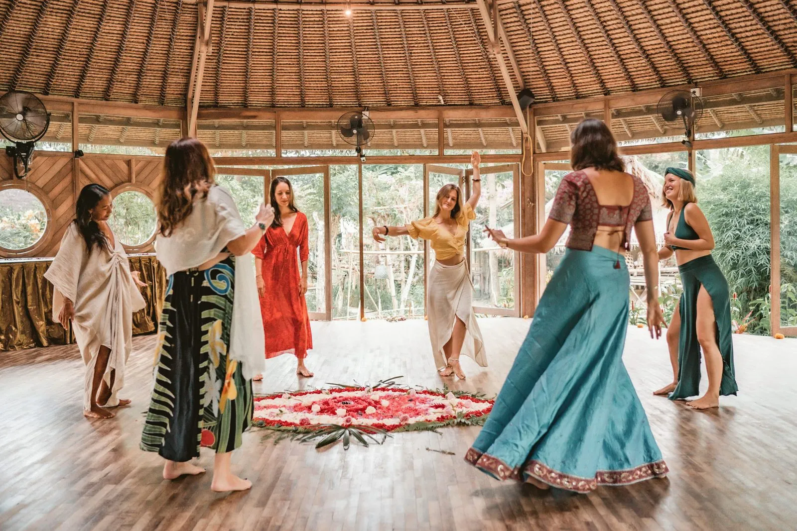Women in long skirts dancing around a circle of petals inside a round bamboo shala