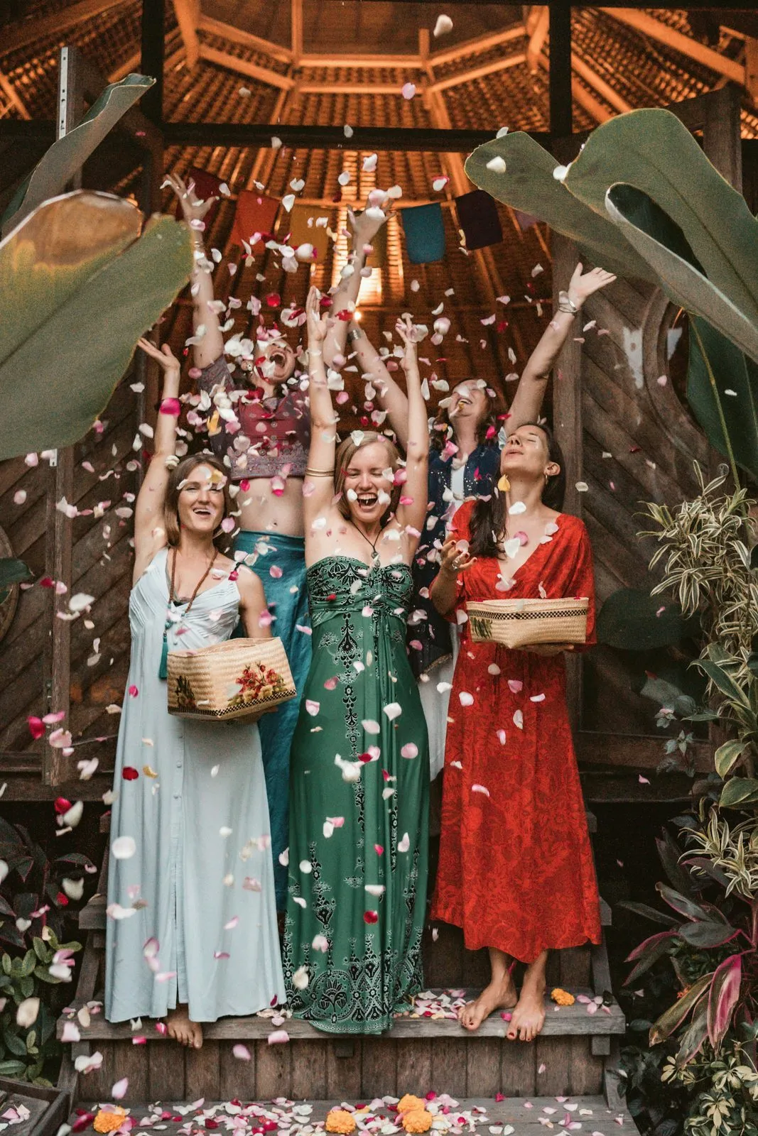 Three women in long dresses throwing petals into the air under a thatched roof, laughing