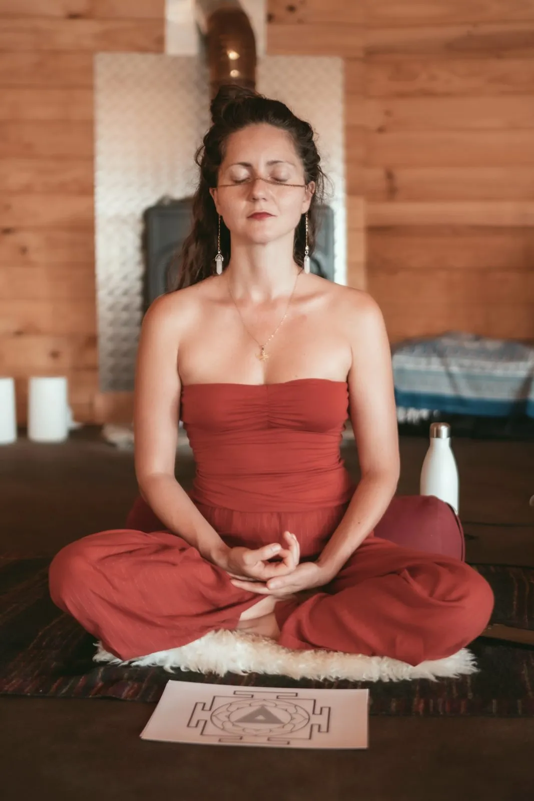 A woman in rust sitting in meditation on a sheepskin in a wooden room, eyes closed