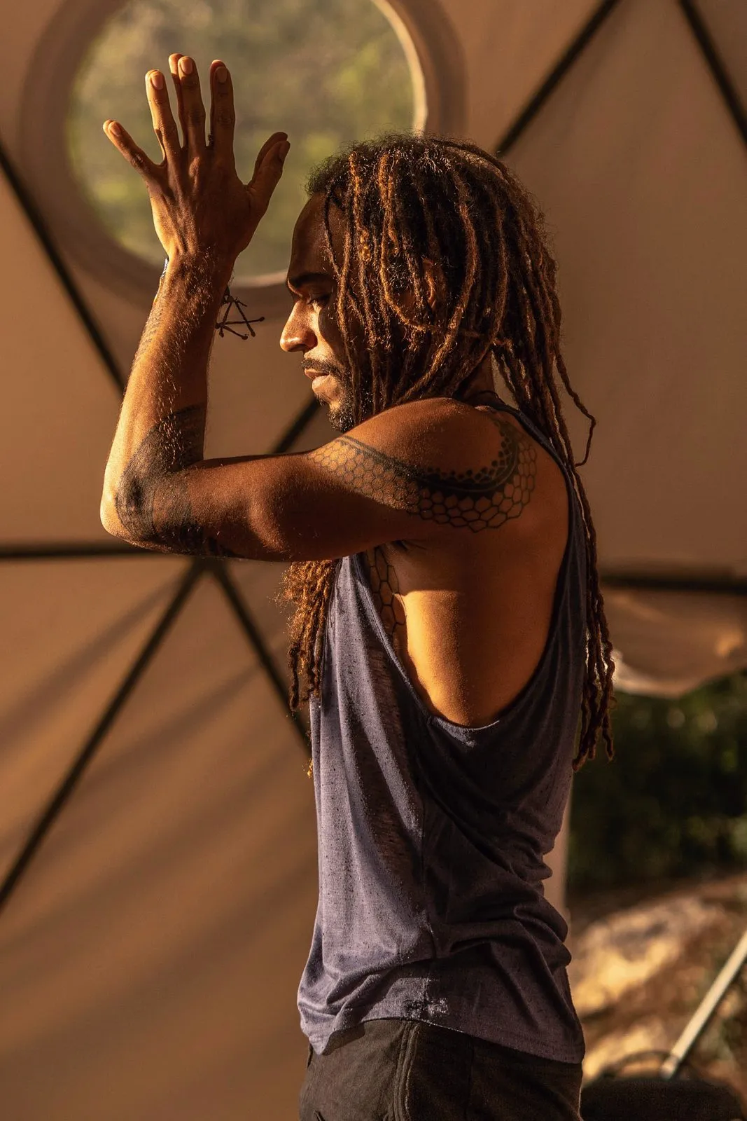 A man with dreadlocks standing with his hands raised in a mudra inside a dome, lit from the side