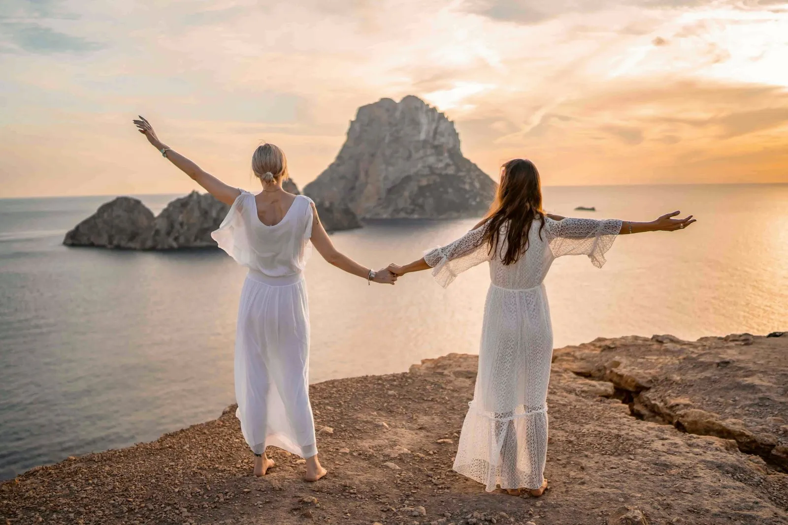 Two women in white holding hands on a cliff edge at sunset, Es Vedrà rising from the sea before them, Ibiza