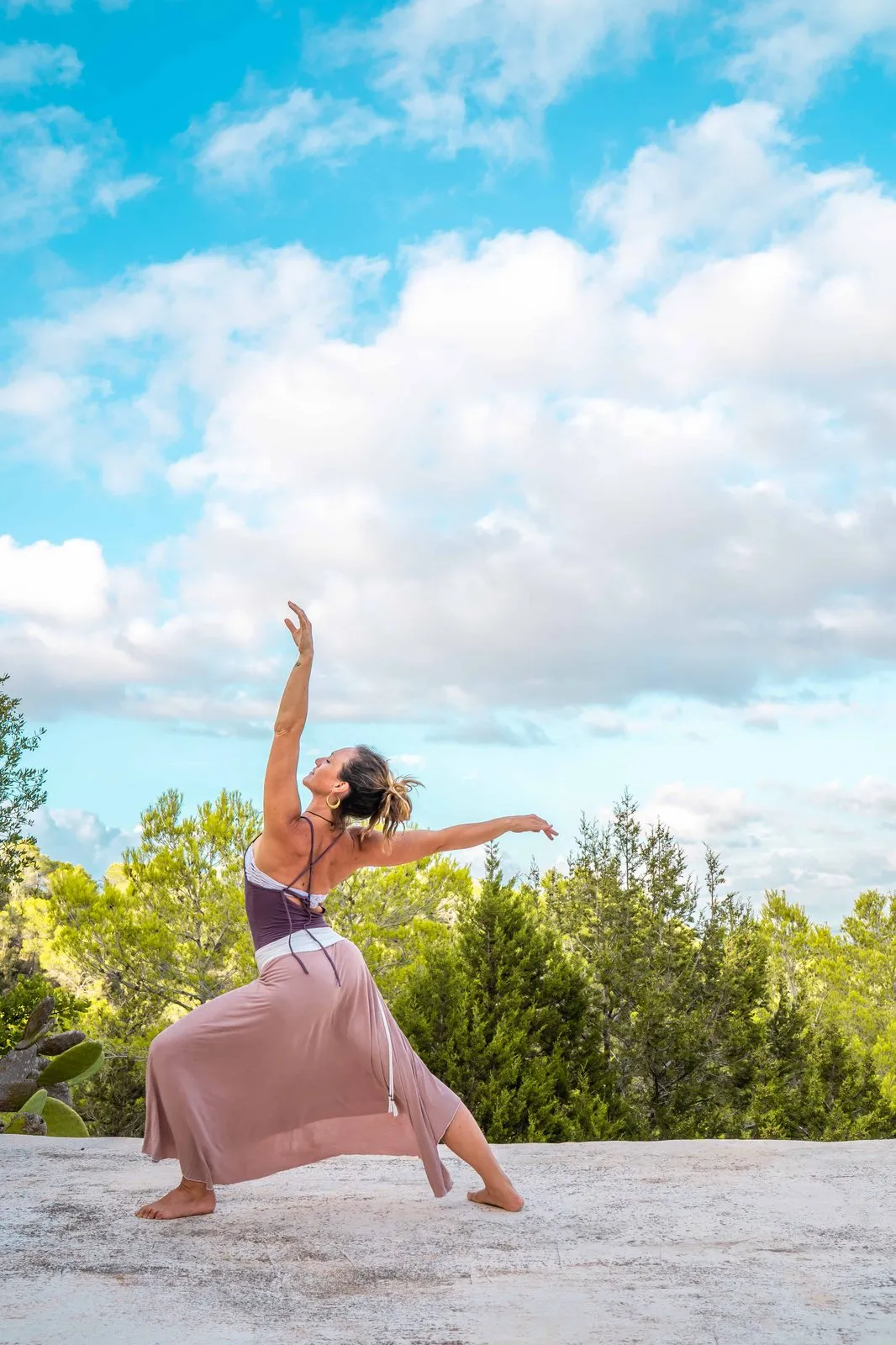 A woman in a dusty pink skirt dancing on a white rooftop under a big blue sky, pines behind her, Ibiza