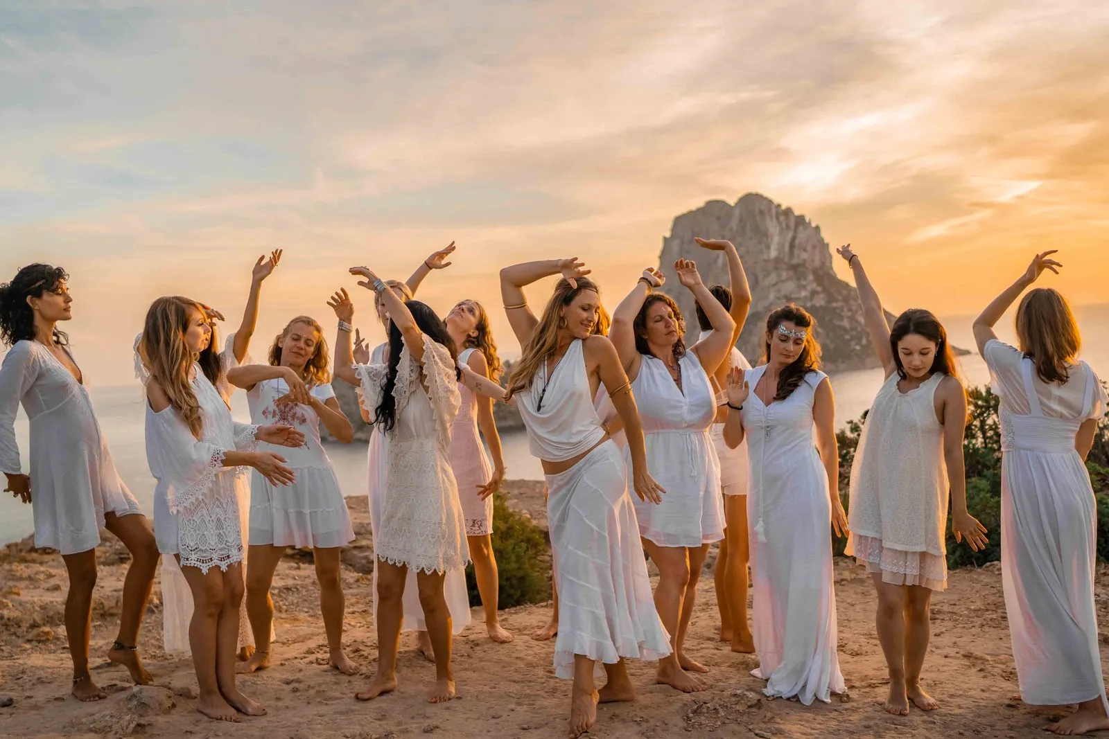 A circle of women in white dancing with their arms raised on a hilltop at sunset, on retreat