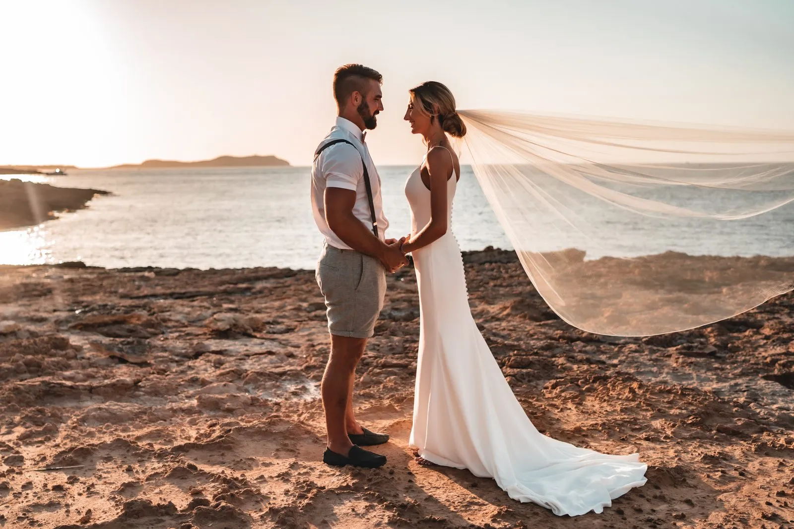 A bride and groom holding hands on the rocks at the edge of the sea at sunset, her veil trailing behind her