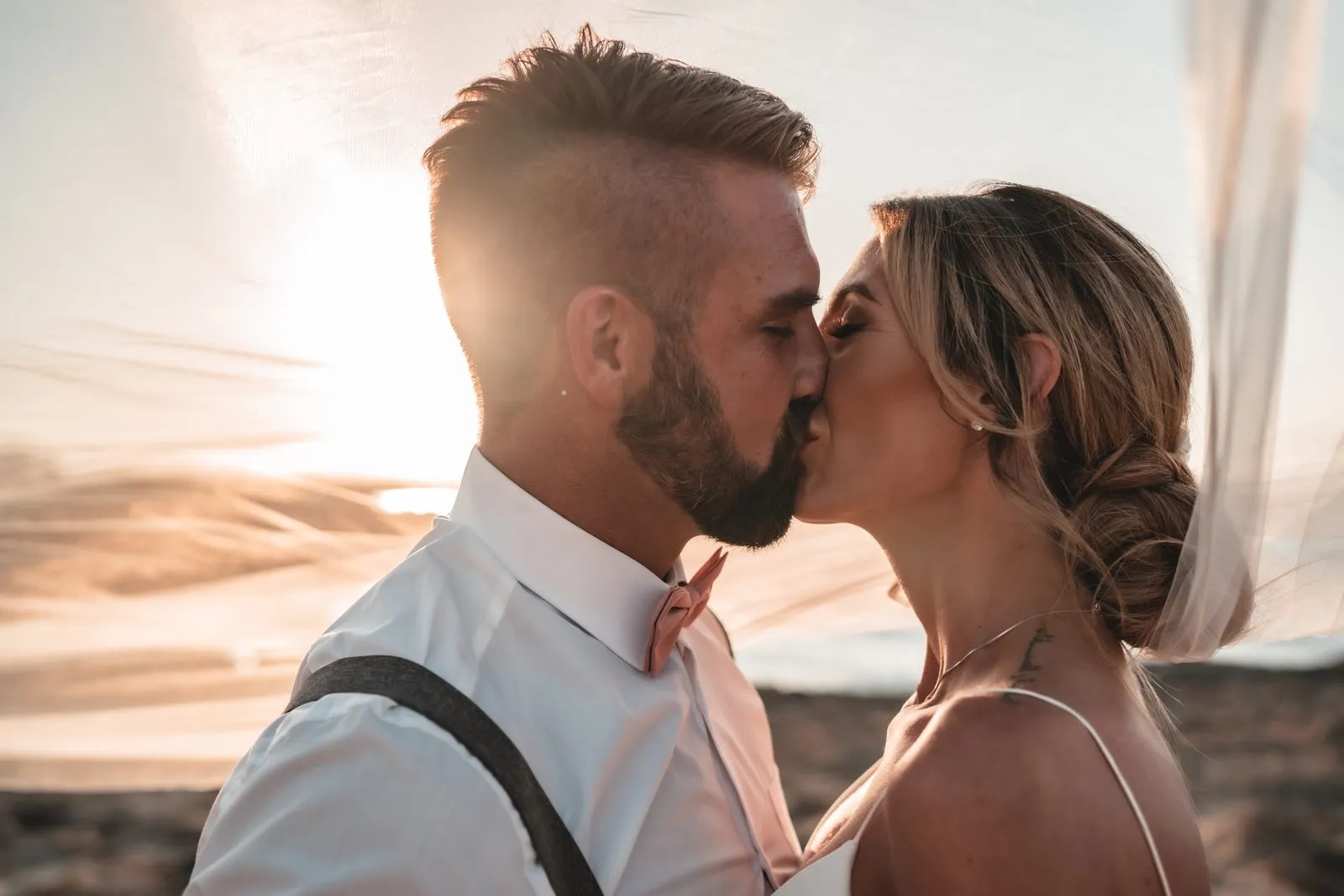 A groom in braces and a bow tie kissing his bride under her veil, the sun setting behind them