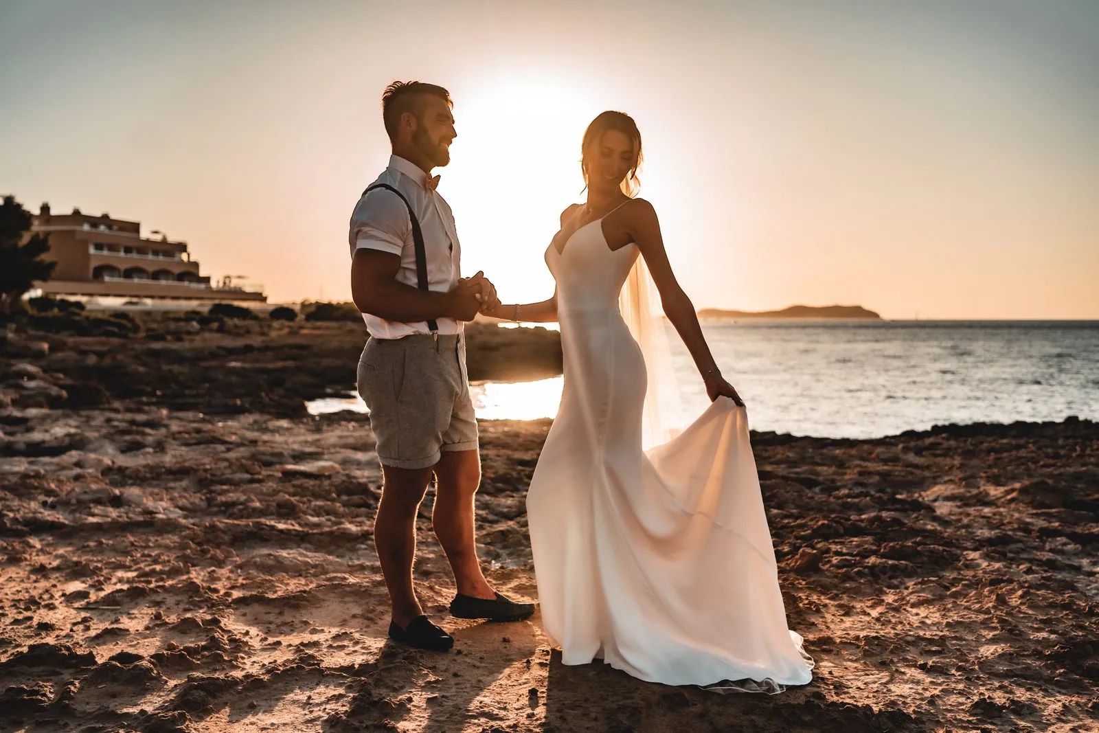 A bride lifting her dress as she and the groom cross the rocks at sunset, the sea behind them