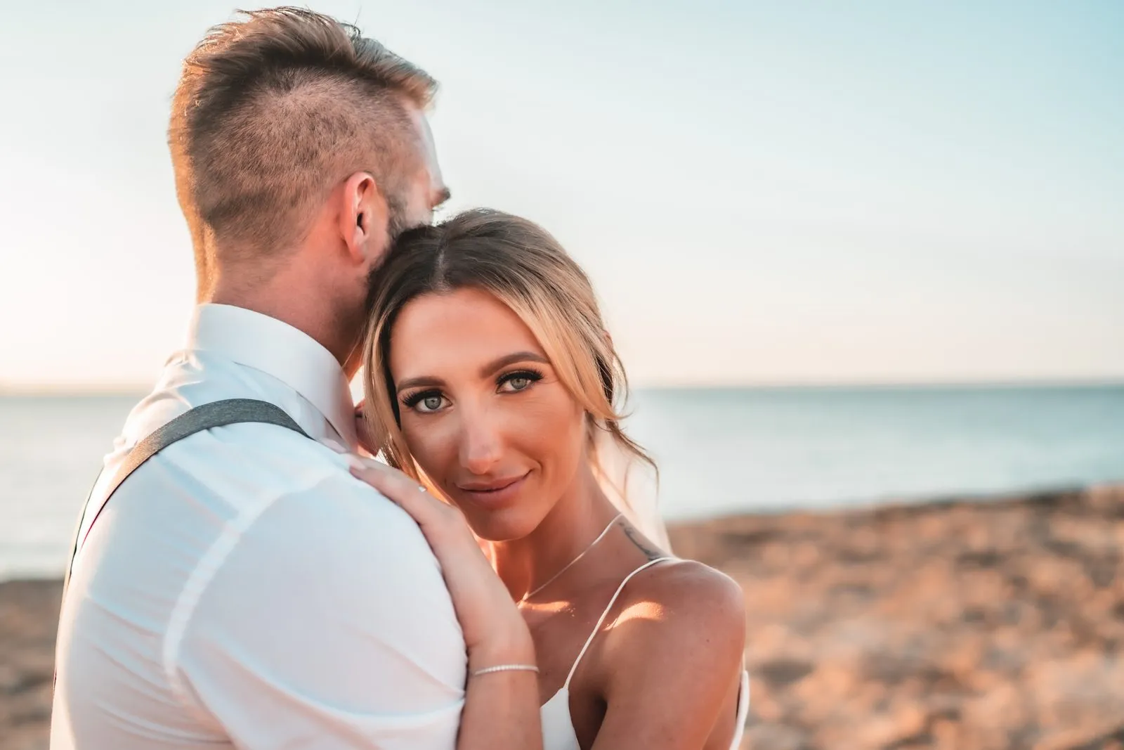 A bride resting her cheek against the groom's chest on the shore, looking to camera