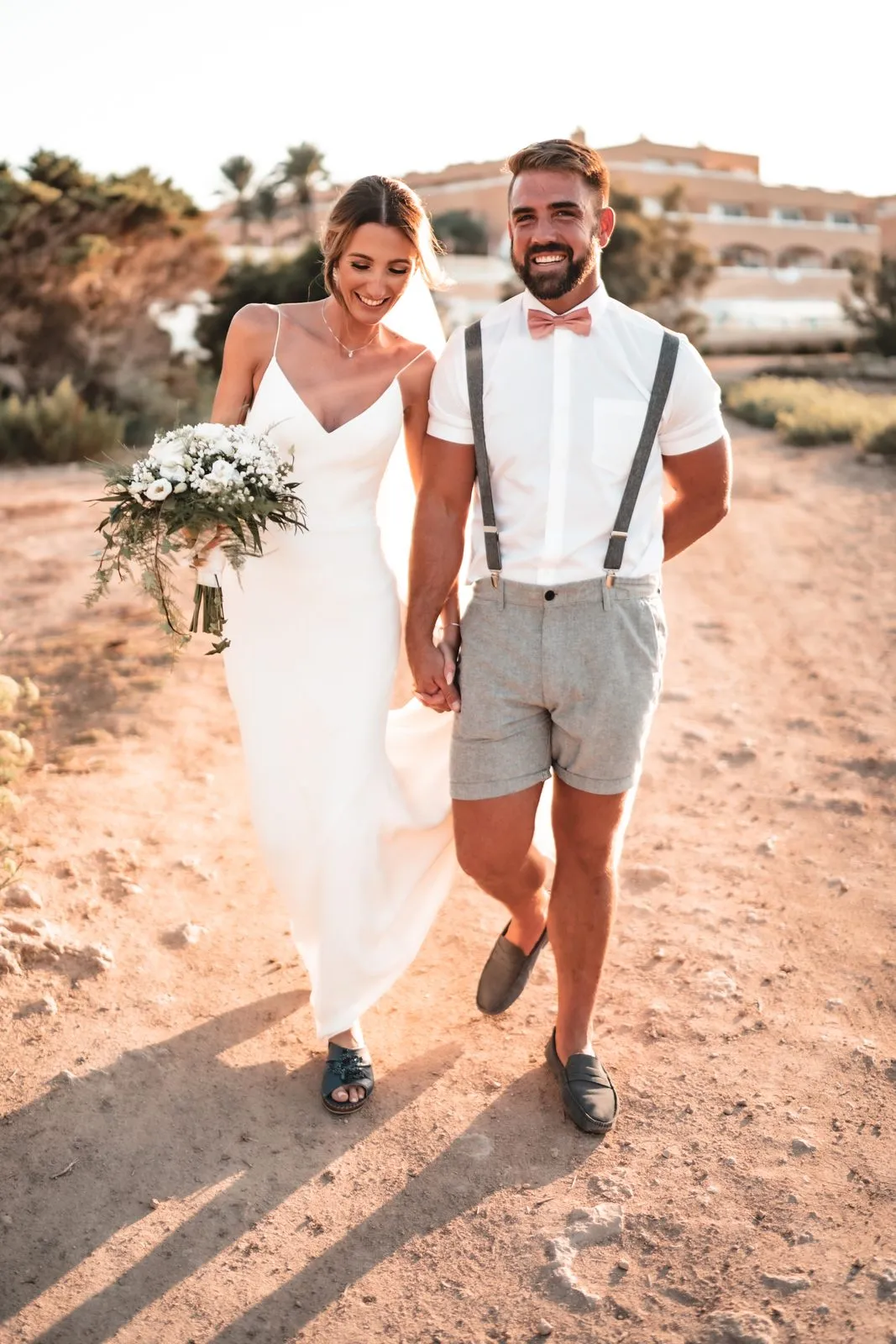 A bride with a white bouquet and a groom in shorts and braces walking a dusty path, laughing