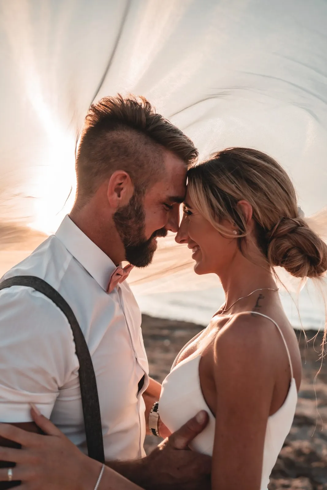 A bride and groom forehead to forehead as her veil blows over them at sunset