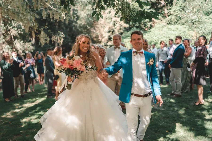 A bride in a full white gown and a groom in a blue jacket walking back down the aisle through their guests in a garden
