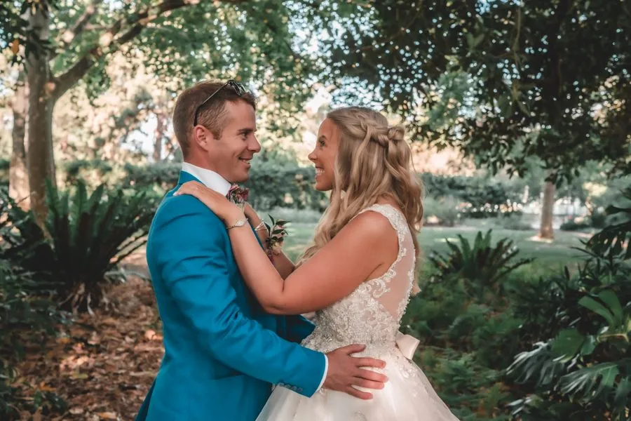 A bride and groom in a blue suit laughing together under the trees of a garden