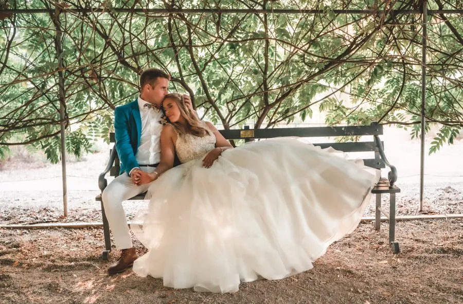 A bride and groom sitting on a bench under a wisteria arbour, her gown spread across the seat