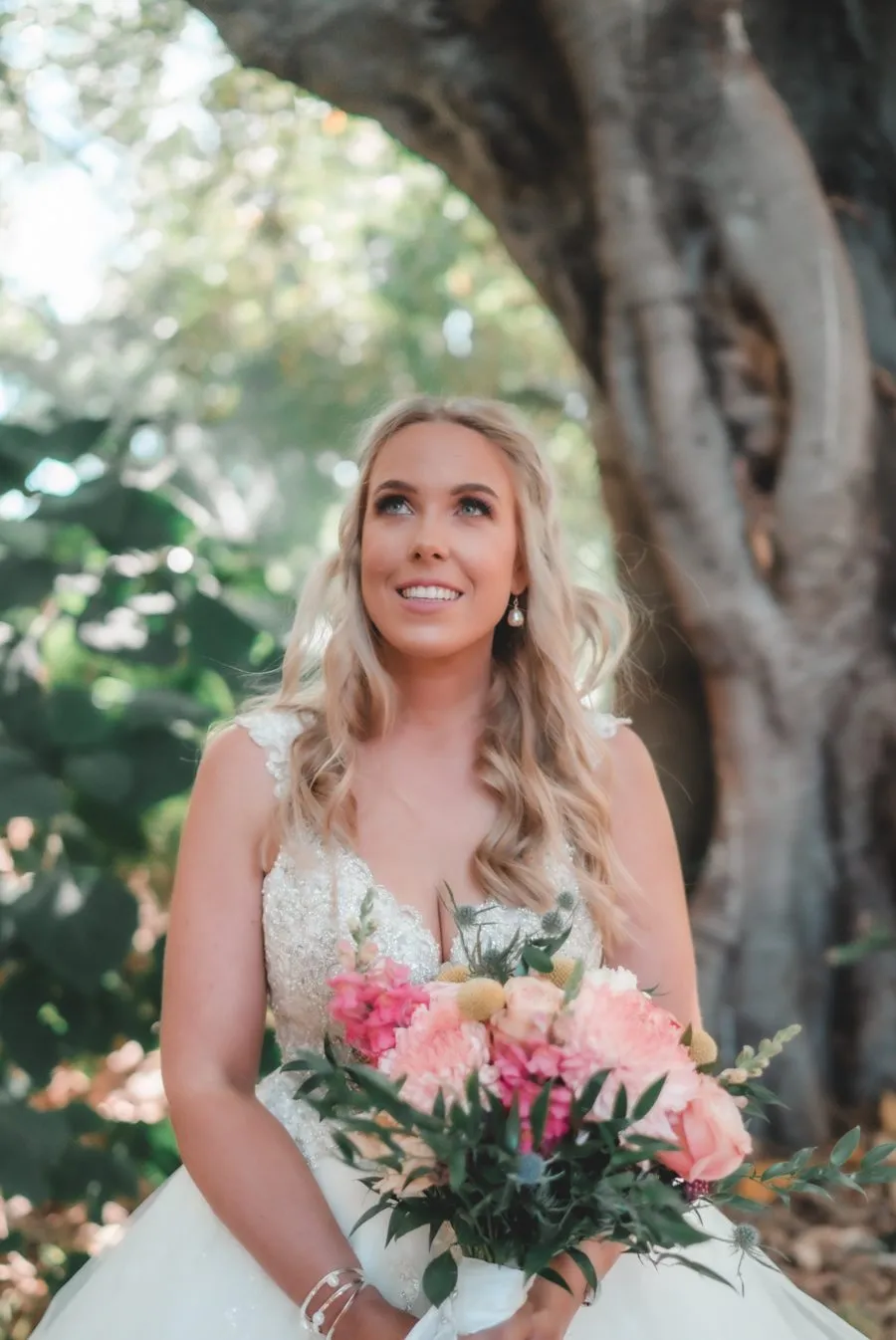 A bride with a pink and white bouquet looking up beneath the boughs of a fig tree