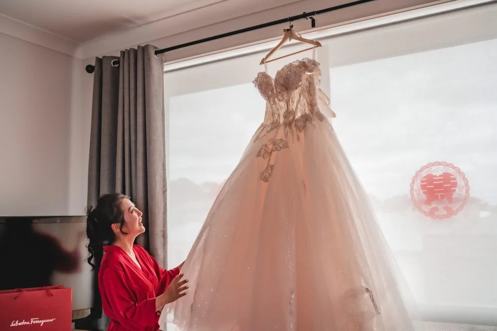A bride in a red robe looking up at her wedding gown hanging in the window