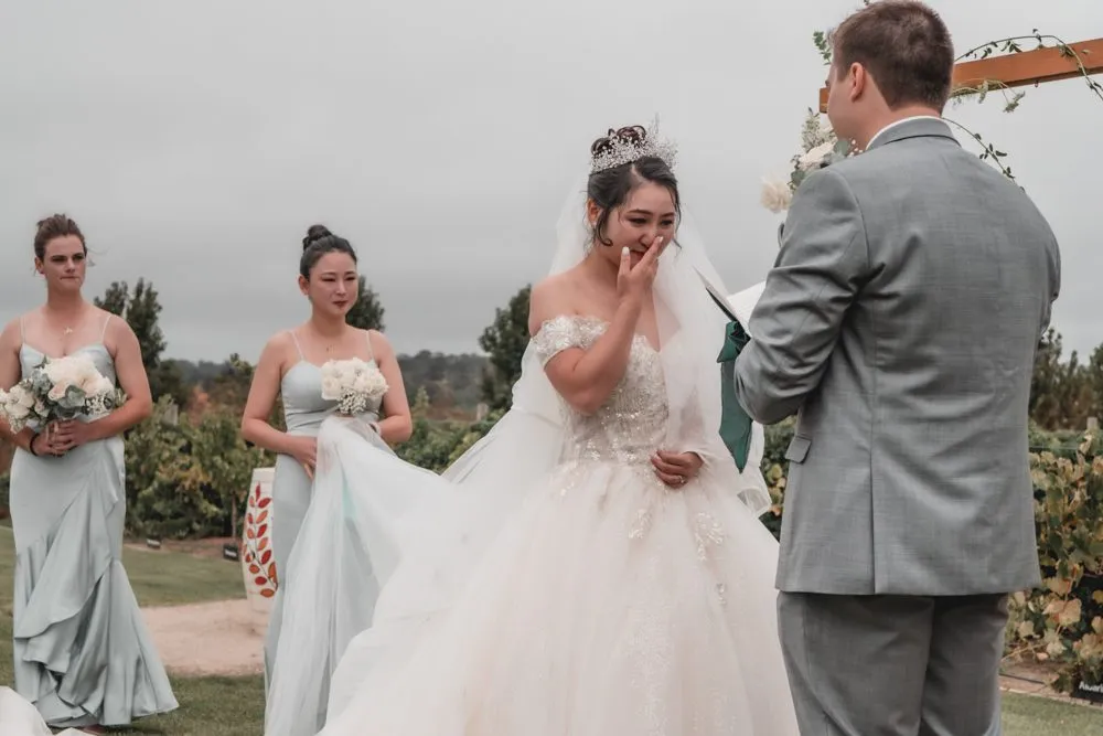 A bride in a tiara wiping a tear during an outdoor ceremony, her bridesmaids in sage green beside her